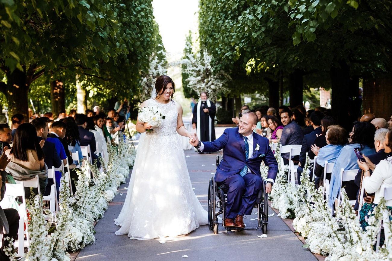 A bride and a groom in a wheel chair walk down the aisle. the aisle is lined with white florals and trees on the outside perimeter. 