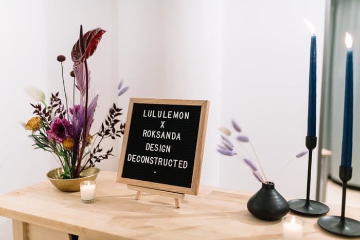 a sparse flower arrangement next to a letter sign with two pillar candles atop a light wooden table