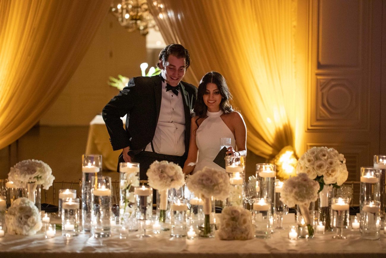 A bride and groom stand at the table admiring the centerpieces and decor. Dramatic up-lit draping is in the background.