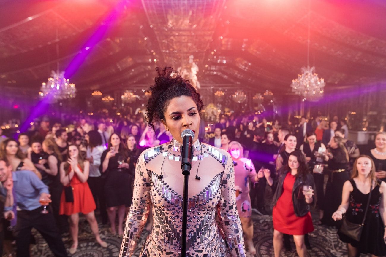 A singer stands on a stage singing with her back to the audience. chandeliers and pink lighting are in the background.
