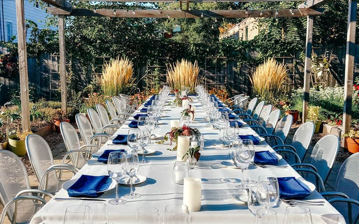 A long table with transparent chairs on either side. A wooden pergola surrounds it with pampas grass and low pillar candles. 