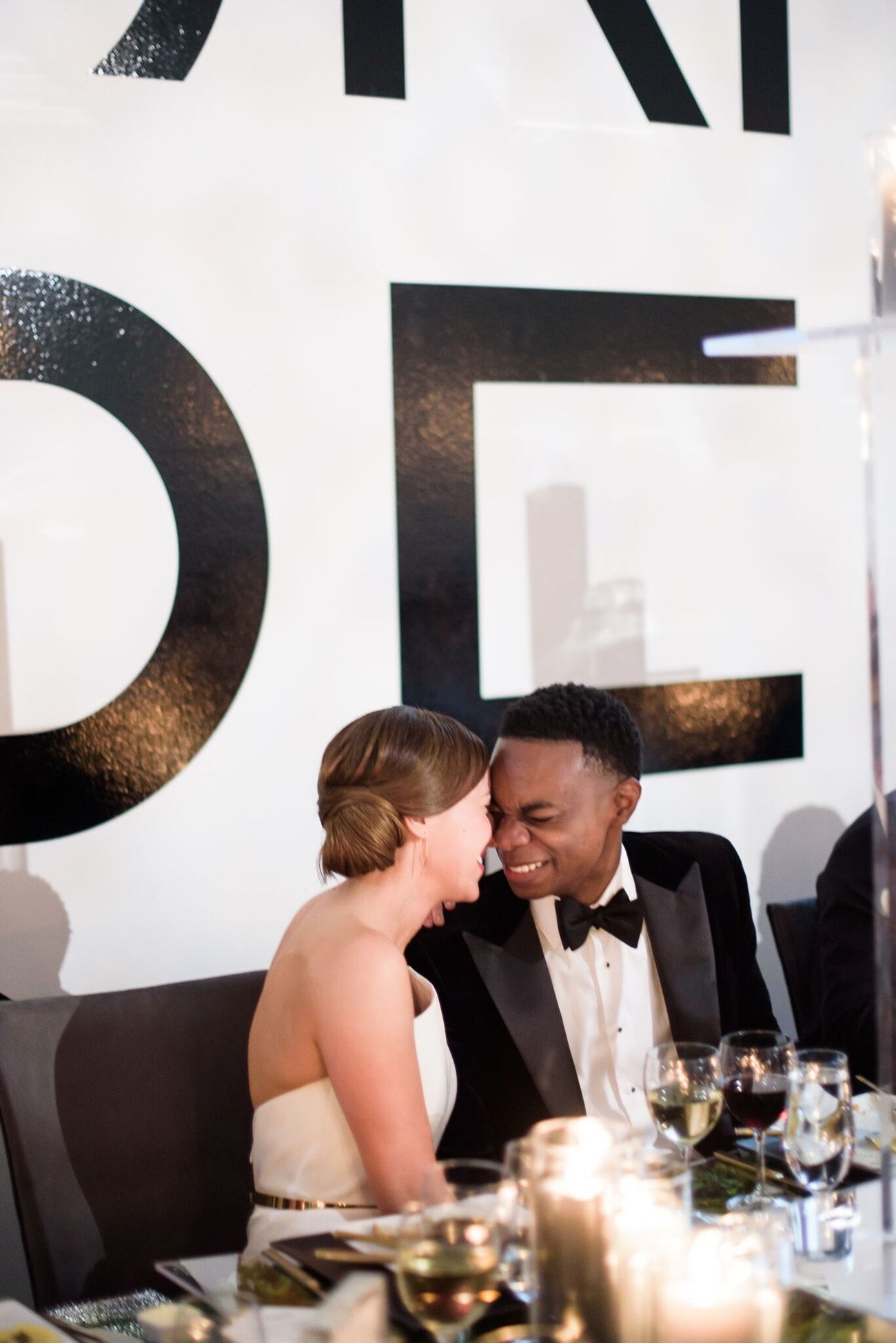 A bride and groom sit at their table at the reception. They are leaning into each other touching faces.