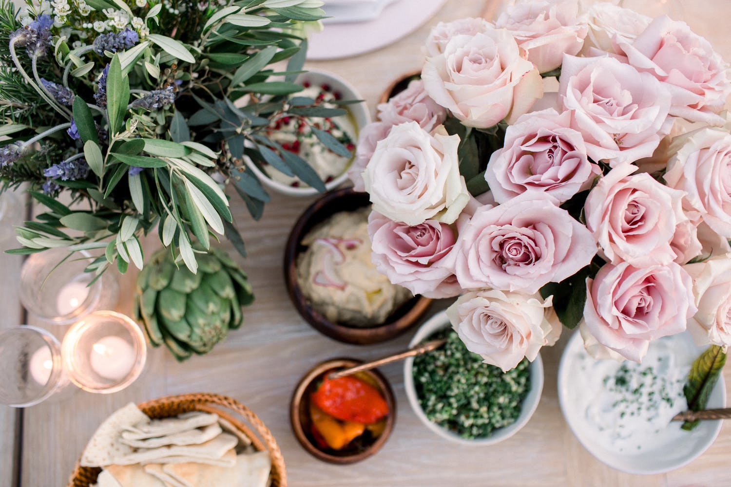 still life photograph of blush-colored roses, sage, artichoke, votive candles, and bowls of pita and various spreads