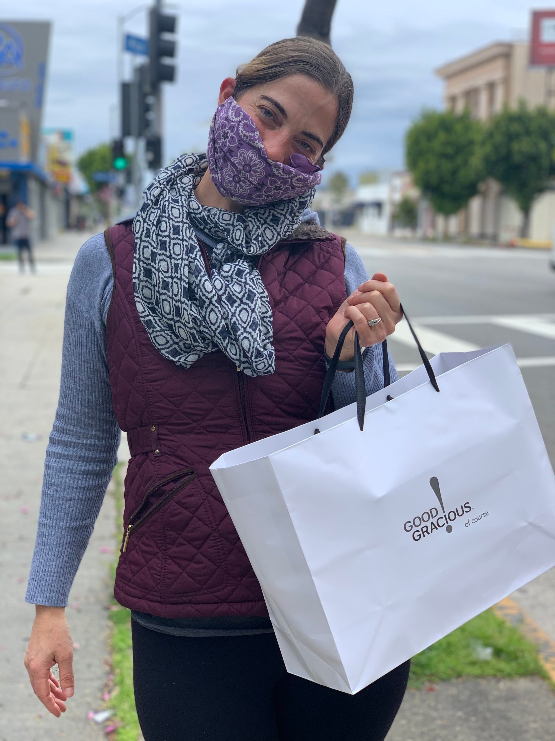 A woman wearing a scard and a face mask holds a white handle bag in one hand white standing on the sidewalk.