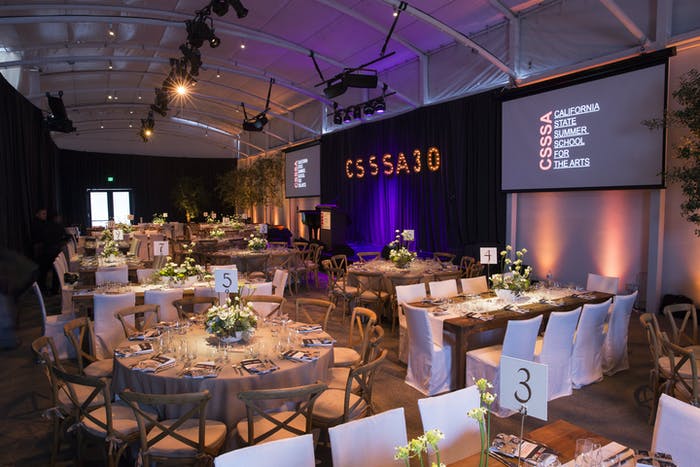 Round tanles surrounding other rectangular tables. There is spotlighting throughout the space and purple lighting next to a screen
