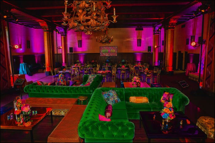 A space that is washed in a coat of pink lighting with a chandelier and green retro couches underneath