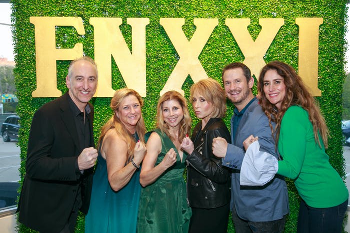 Six men and women stand in front of an photo backdrop with grass like background and yellow lettering