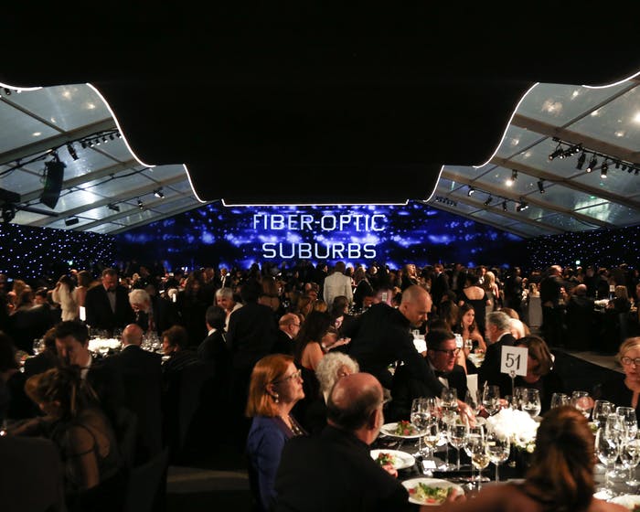 An outdoor event is flanked by two ceilings that don't meet in the middle. A screen is in the center of the room and people congregate
