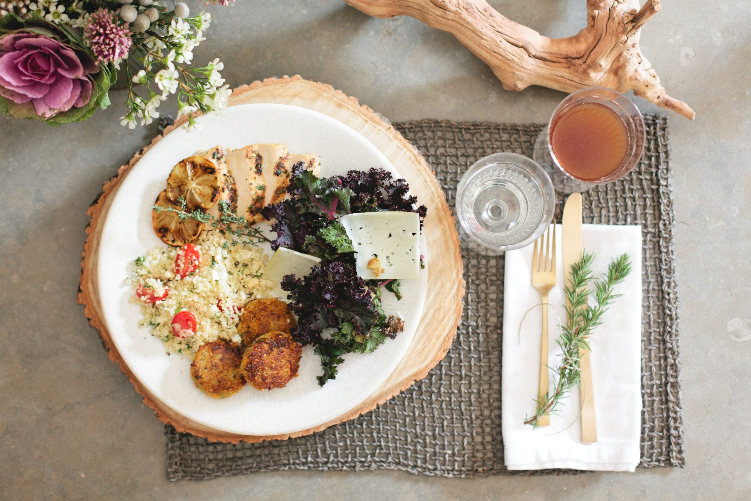 A white plate of artfully catered food rests on a circular wooden placemat atop a gray woven square placemat. White blooms and a driftwood branch accompany a glass of water, a glass of cider, and a white napkin with gold utensils and a sprig of greenery.