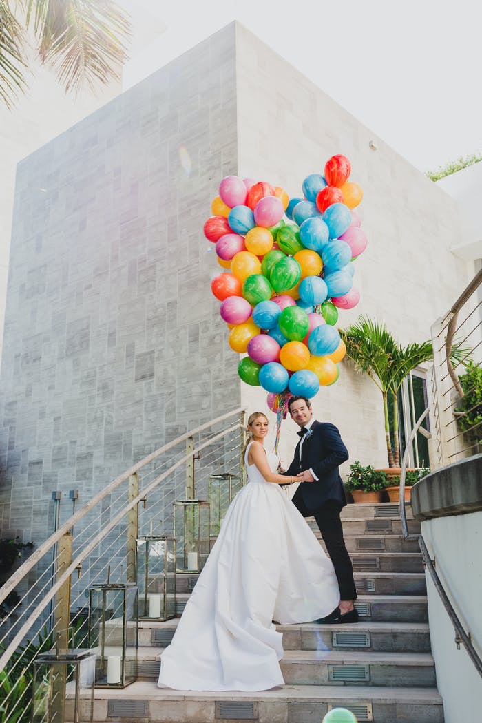 A couple posing on a winding staircase with a balloon bouquet mad of pink, blue, green, and yellow balloons