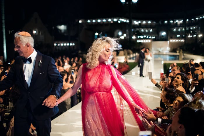 A mom in a fashionable red dress pauses on the aisle of her her child's evening wedding ceremony to greet guests against a backdrop of lit-up buildings.