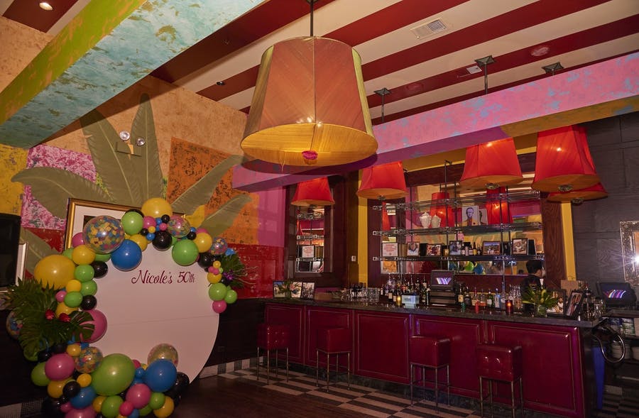 A dim bar on the right with bottles along the inside. A photo backdrop to the left with a multicolor balloon border.