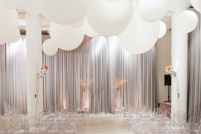 A white room with wide white columns and a white curtain. White decor sits in front of the curtain and large circular balloons float to the ceiling