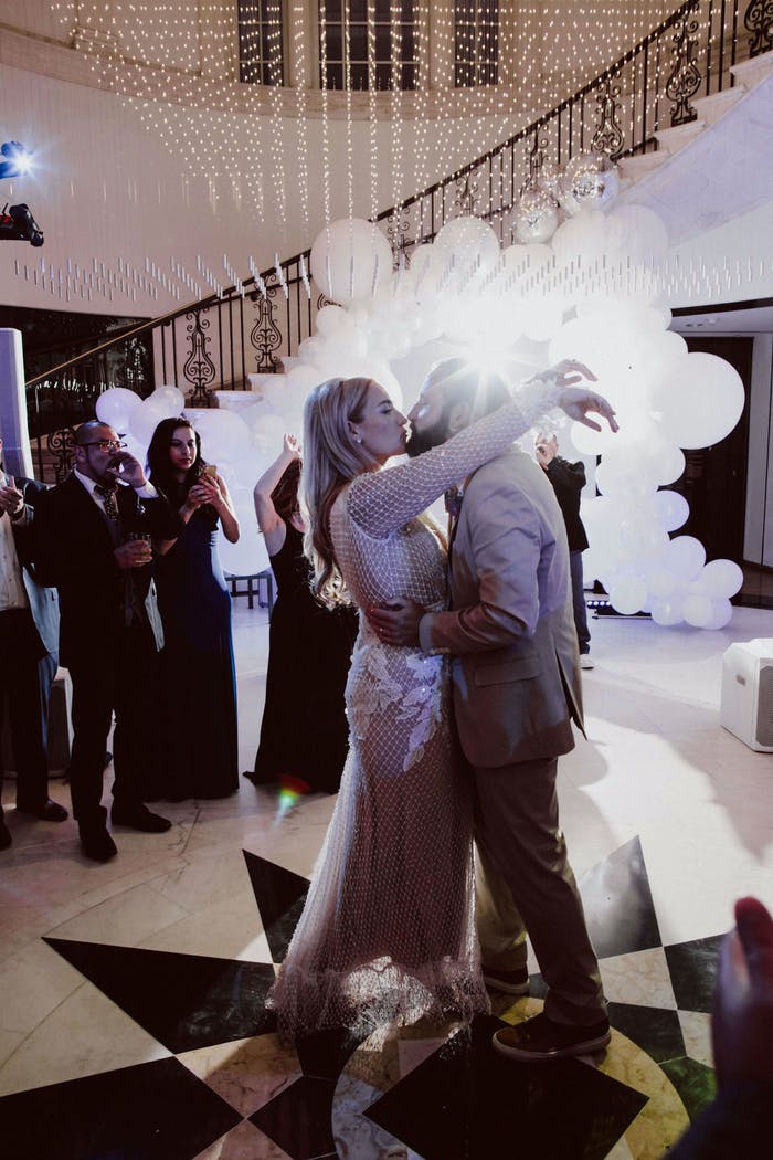 A couple standing in front of a white balloon arch and a crowd of people. A winding staircase is behind them