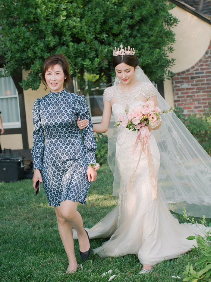 A mom leads her daughter to the aisle wearing a silver dress. There is grass and a tree behind them