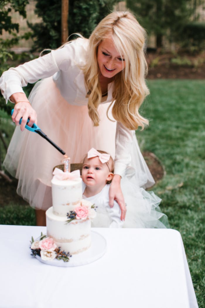 Outside, a mom wearing a long-sleeved white shirt and soft pink frilly skirt leans over her young child wearing a pink bow and lights a white two-tiered birthday cake with pink roses.