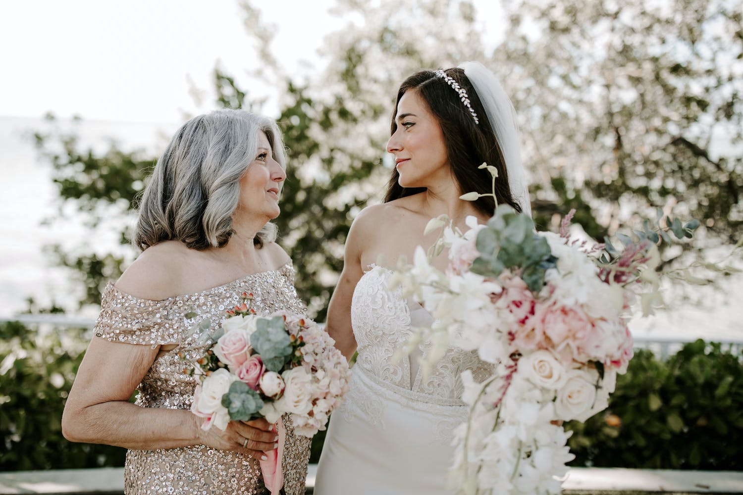 Close up of bride sharing a meaningful look with her mother. Both hold blush and white bouquets with greenery.