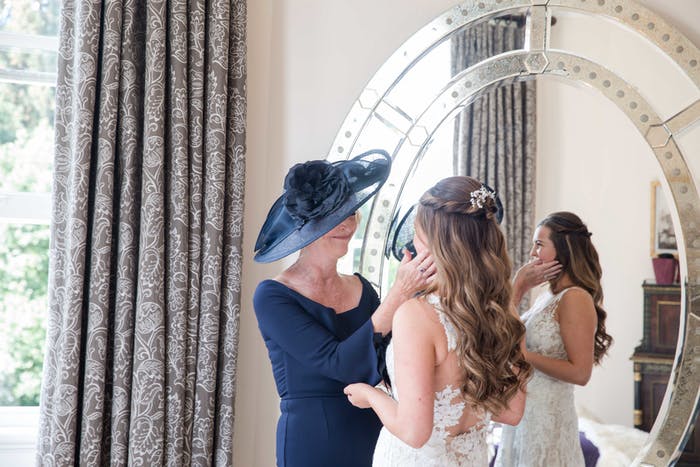 A mother wearing a large brimmed hat stands in front of a mirror with her daughter to put on her necklace