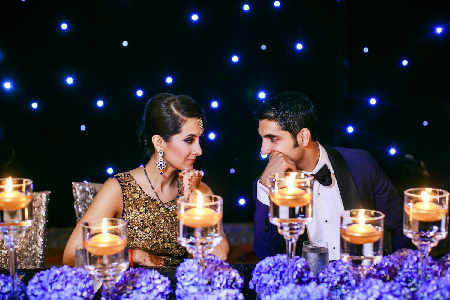 Bride and Groom Sit at Sweetheart Table Laden With Purple Hydrangea Wedding Centerpieces and Candlelight Against a Starlit Backdrop | PartySlate