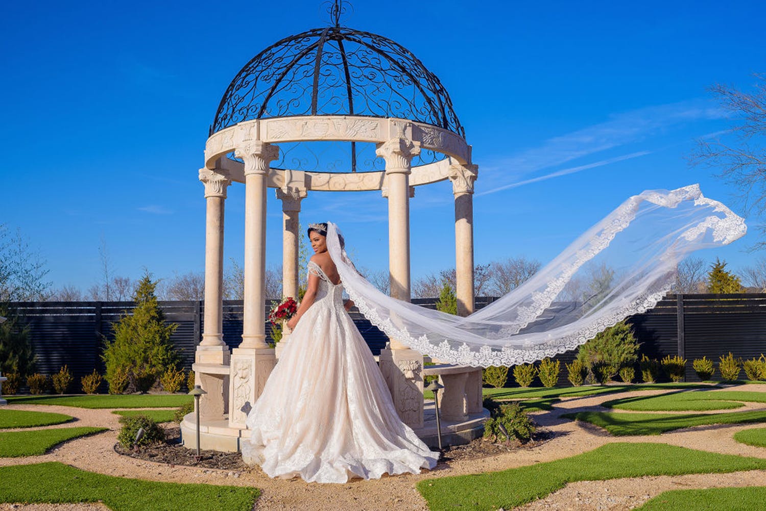 Bride with blowing veil stands in front of pillared wedding arch with filigree dome | PartySlate
