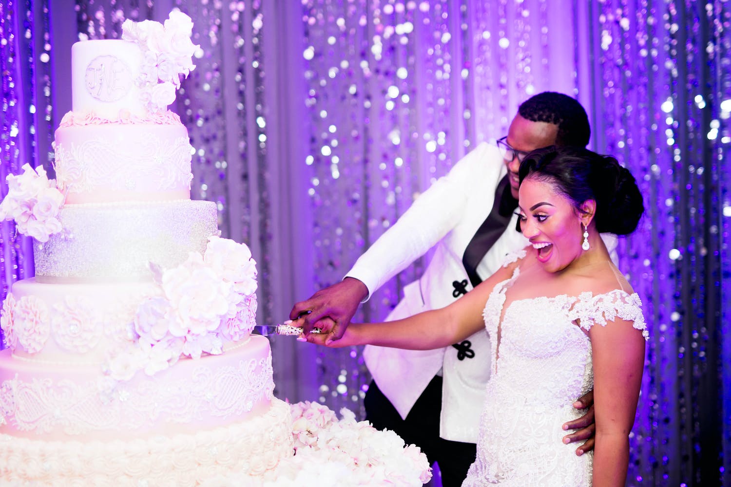 Bride and Groom Cut into Towering White Wedding Cake Against a Glittery Purple Backdrop | PartySlate