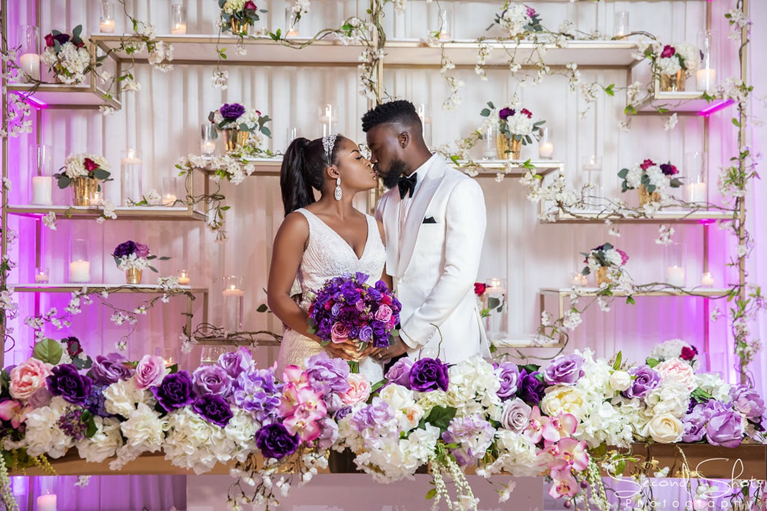 Bride and groom kiss in front of glamorous purple and gold wedding backdrop | PartySlate