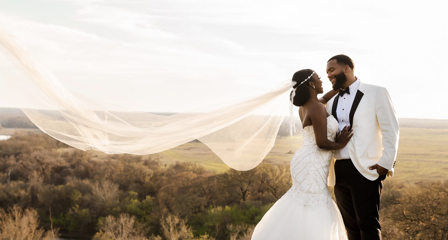 Bride and groom with bride's veil blowing in wind against landscape backdrop | PartySlate