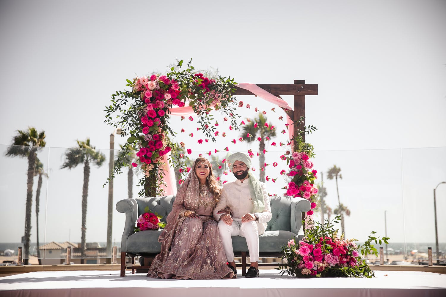 Bride and groom sit on blue settee behind carnation curtain at Indian Wedding Ceremony | PartySlate