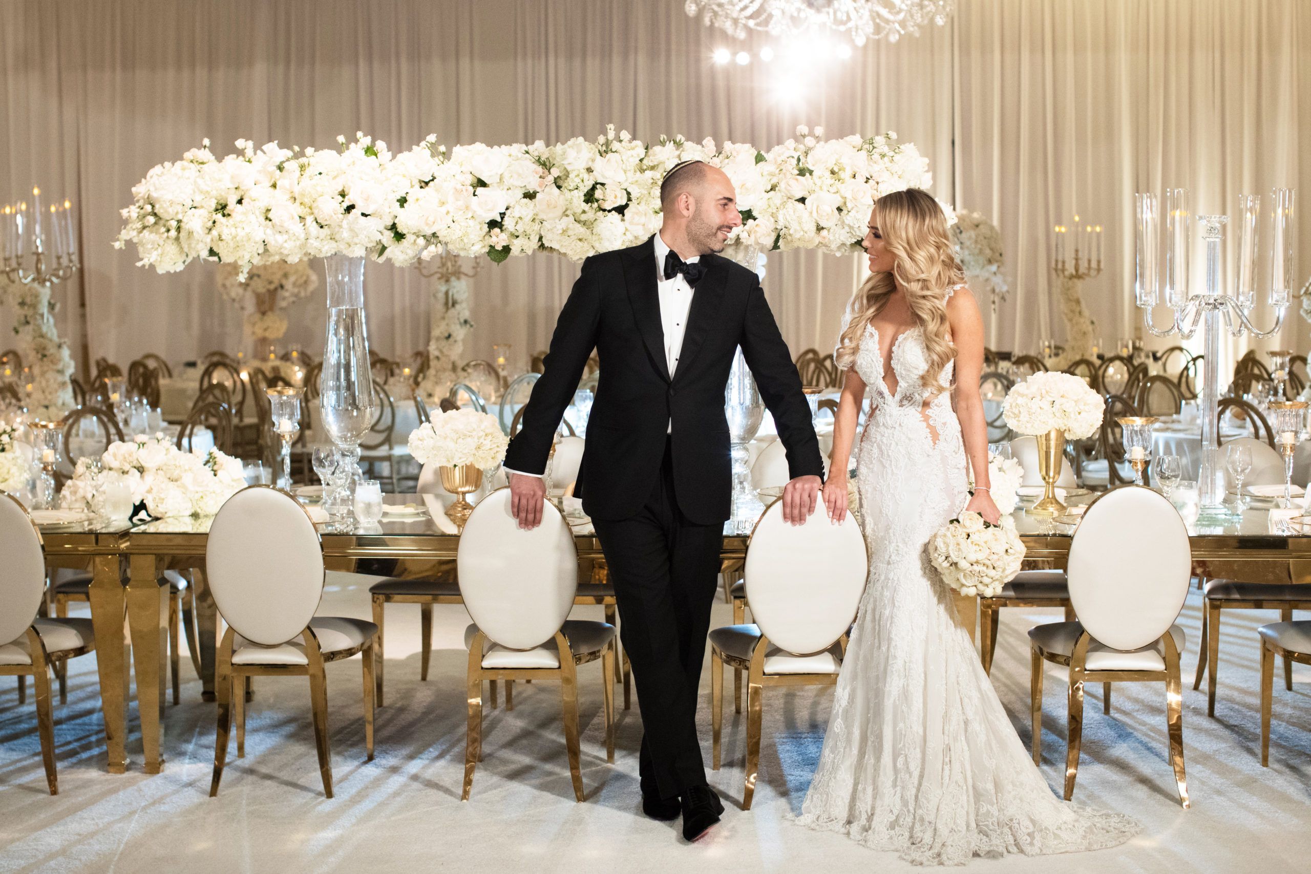 Bride and Groom Standing In Front of Table Filled with White Flowers and Drapery | PartySlate