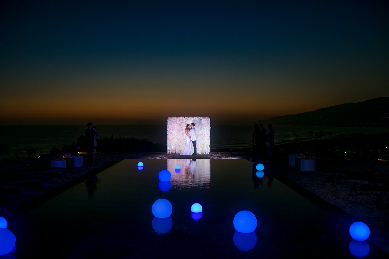 Couple kiss at sunset with pink flower backdrop behind them and pool with illuminated blue orbs in front of them | PartySlate
