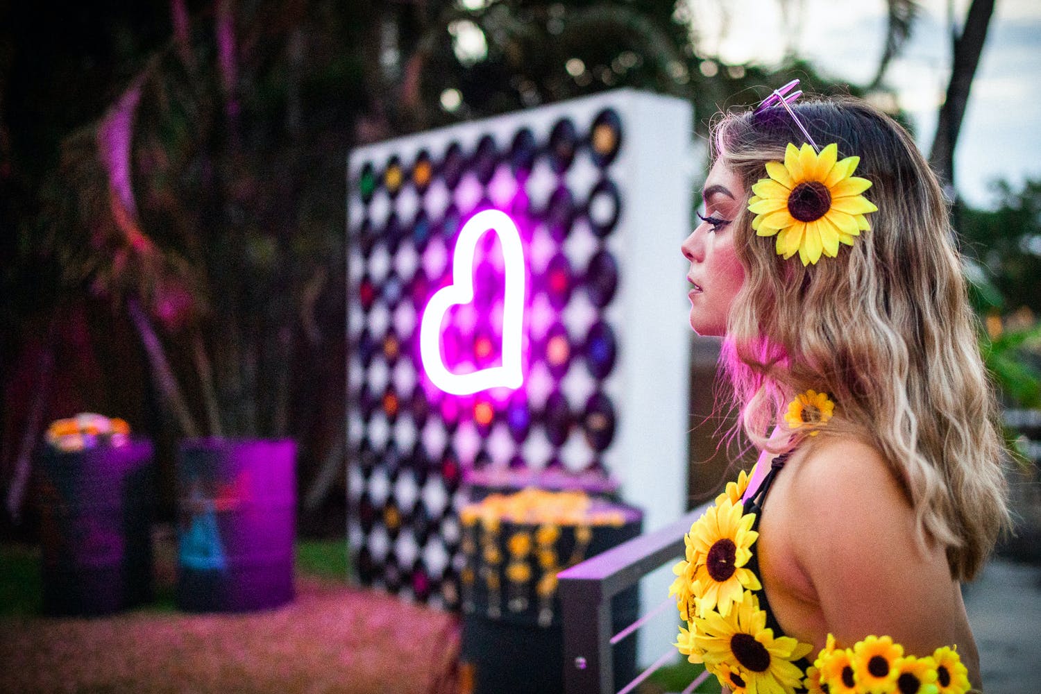Girl with Sunflowers In Her Hair and Dress Standing in Front of Neon Pink Heart Sign | PartySlate