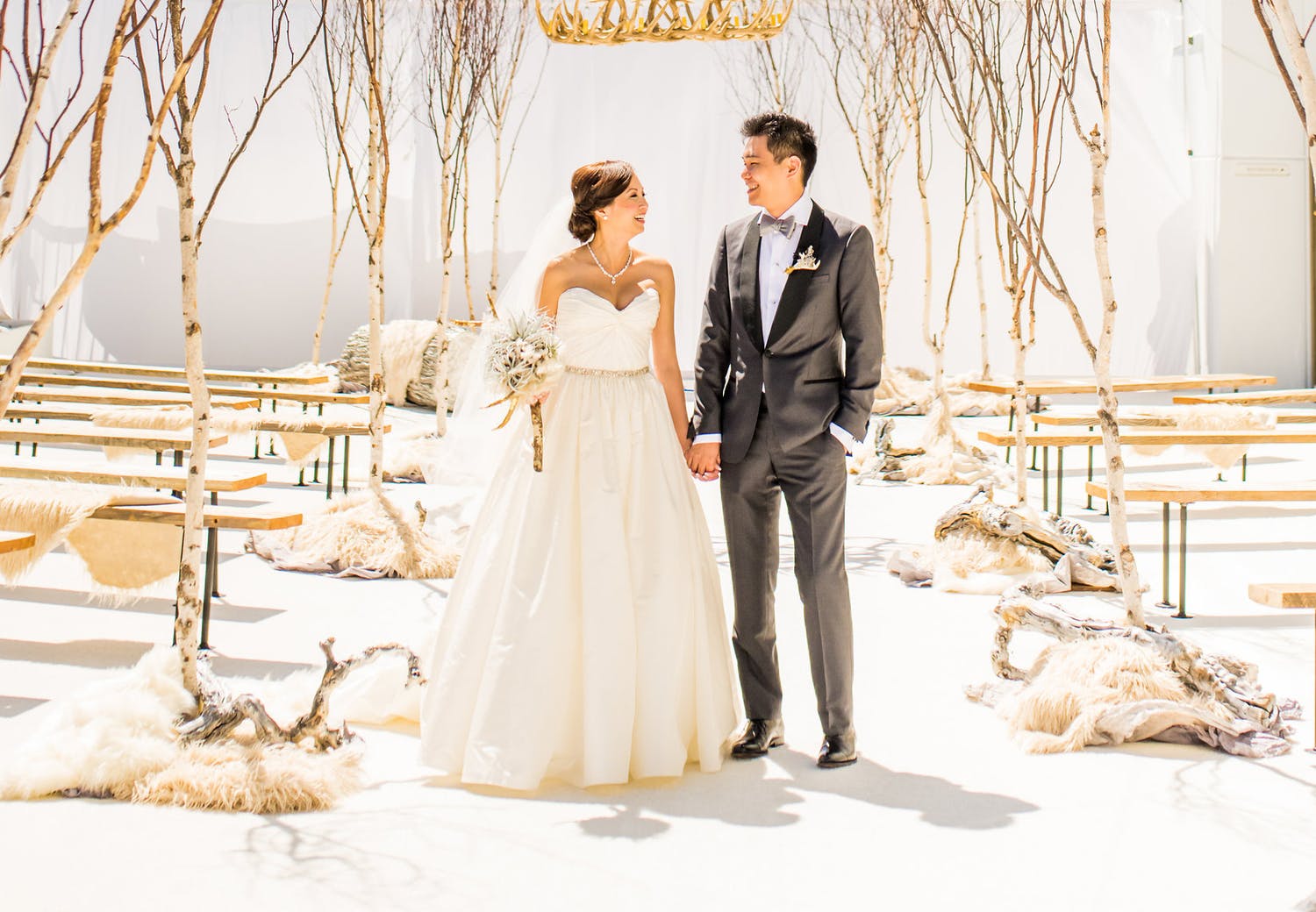 Couple poses in aisle at winter wedding without flowers and soft whites, grays and a natural wood palette as well as bare branch trees | PartySlate