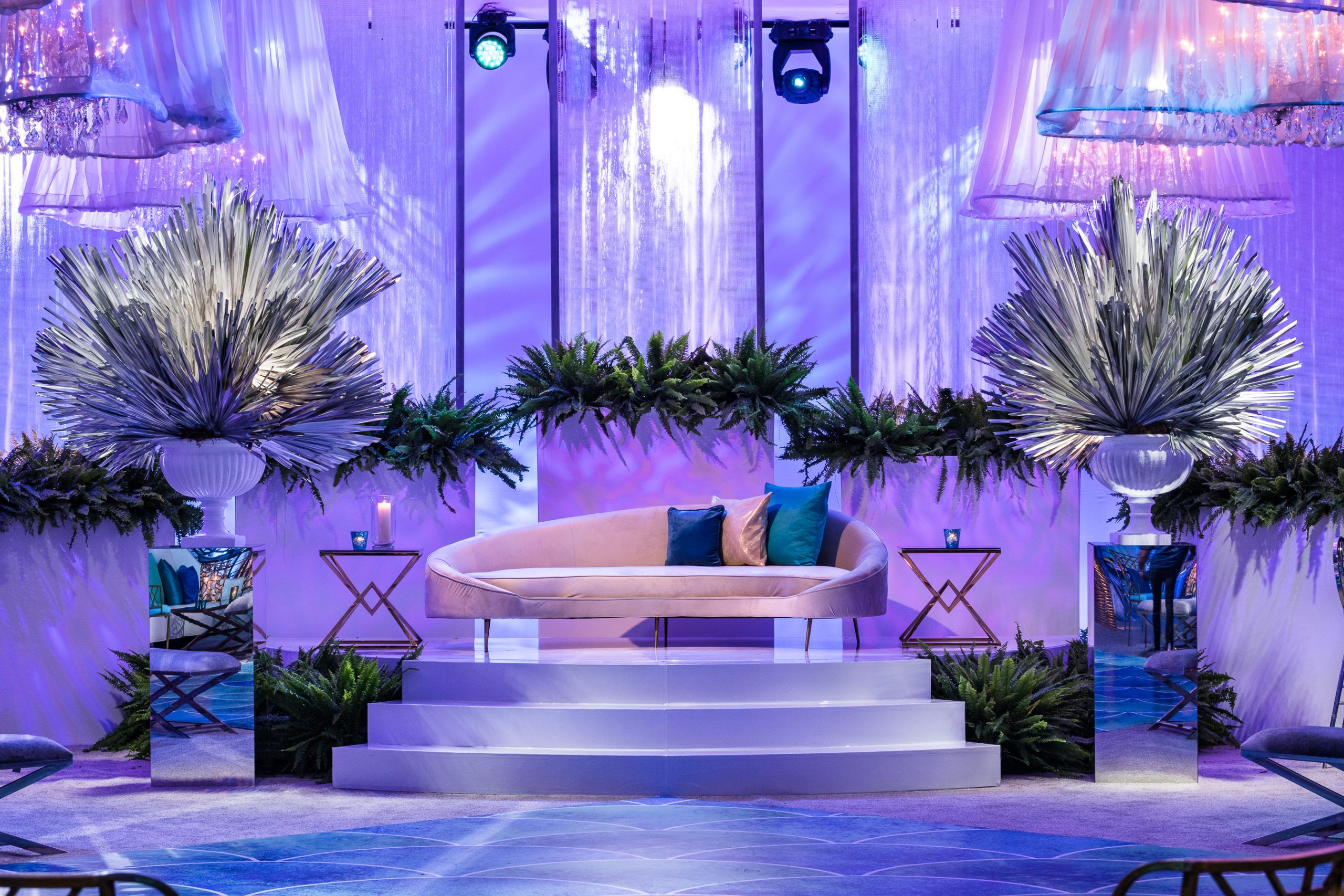A stage lit purple with a white couch and lush greenery