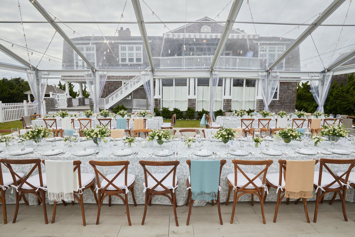 Pretty Hamptons wedding in transparent tent with apricot, white, and light blue wraps thrown over wooden reception chairs | PartySlate