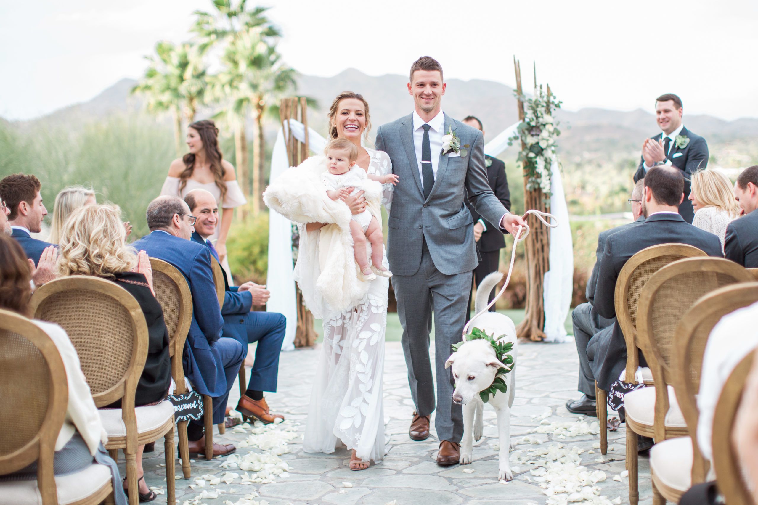 Couple Walking Down Aisle With Baby In Bride's Arms and Groom Walking Dog With Guests Surrounding | PartySlate