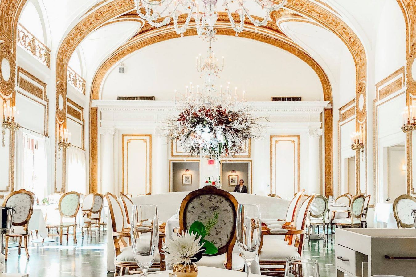 stunning bright private dining area with gold accents and white walls and tables set with floral ceiling installation | PartySlate
