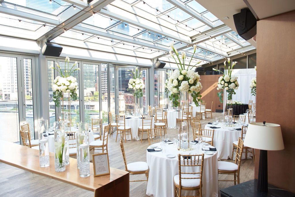 Round banquet tables in reception hall with floor to ceiling windows and white and green floral centerpieces on each table | PartySlate