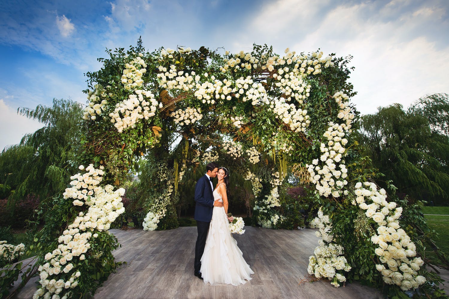 Couple stand beneath wedding arch of lavish greenery and white flowers | PartySlate