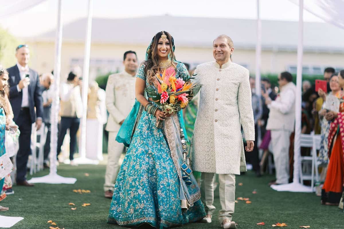 South Asian bride walks down the aisle wearing blue dress and holding tropical bouquet | PartySlate