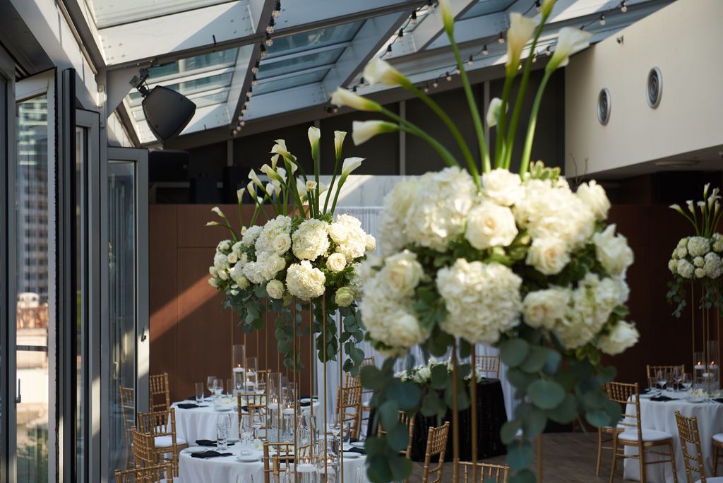 White and green tall centerpieces on round tables in reception hall with large windows | PartySlate