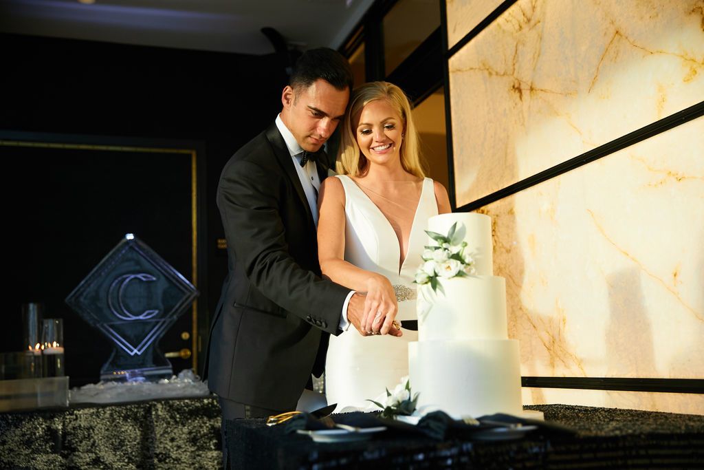 Couple cutting into their white wedding cake with a little bit of greenery on it | PartySlate