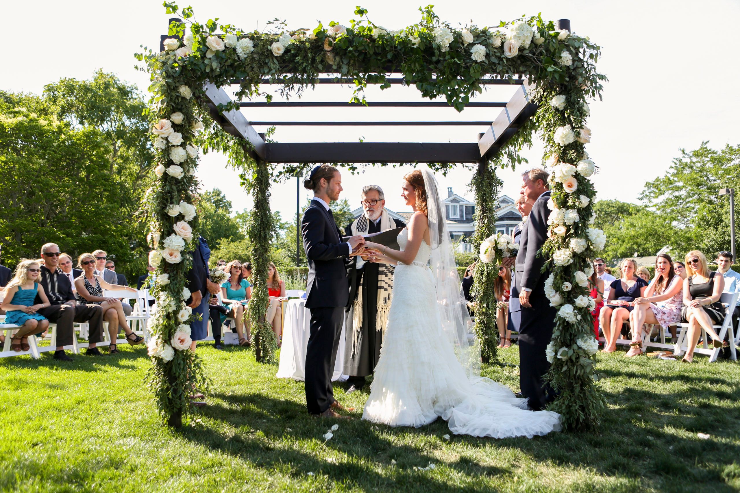 couple stadning at alter getting married with floral arch and seated guests behind them at Harbor View Hotel | PartySlate