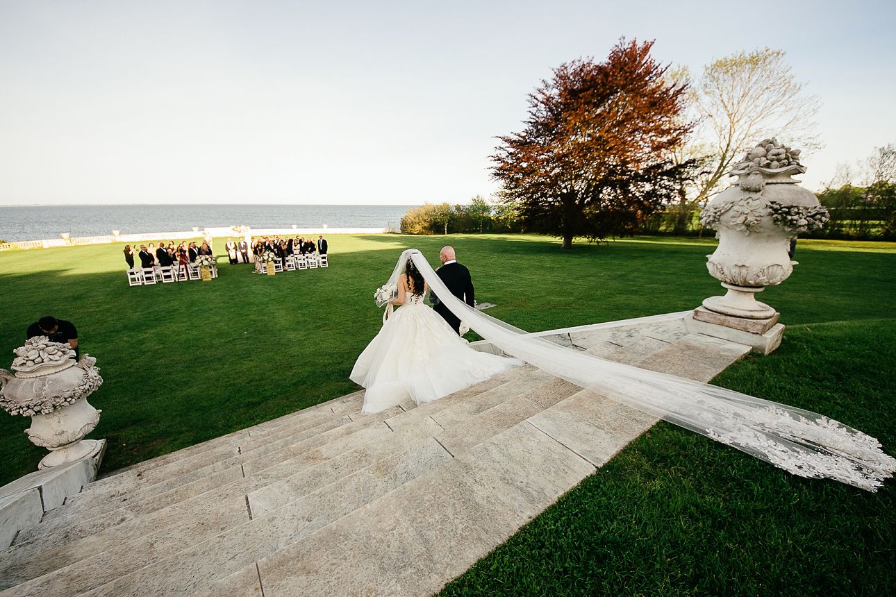 Gilded mansion wedding at Rosecliff Mansion in Newport Rhode Island with bride walking down aisle with long vail train following behind her | PartySlate