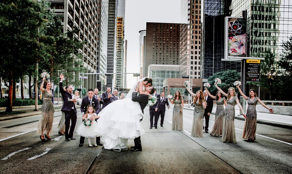 Couples kisses with wedding party and city buildings in background at The Houston Club, a Houston wedding venue.